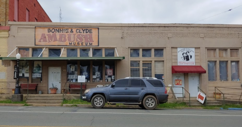 Exterior of the Bonnie & Clyde Ambush Museum, featuring a gray SUV parked outside and a sign indicating it's open.