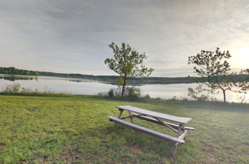 A serene lakeside view with a wooden picnic table and trees, reflecting calm waters under a cloudy sky.
