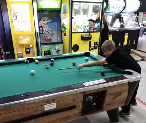 A young boy plays pool at an arcade, focusing on his shot while colorful arcade games are visible in the background.