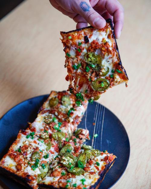 A hand lifts a slice of cheesy pizza topped with jalapeños and herbs from a black plate.