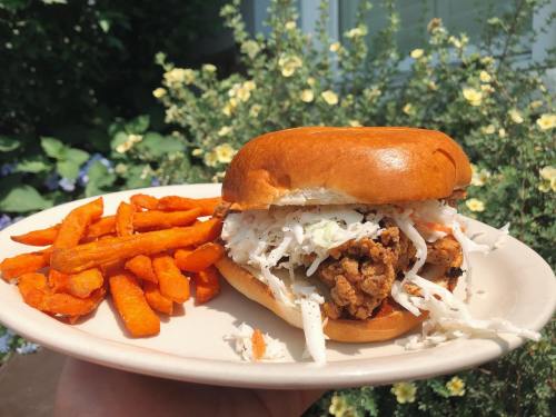 A hand holding a plate with a fried chicken sandwich topped with coleslaw and a side of sweet potato fries.