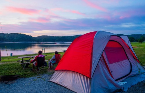 A red and gray tent by a lake at sunset, with two people sitting nearby, enjoying the serene view.