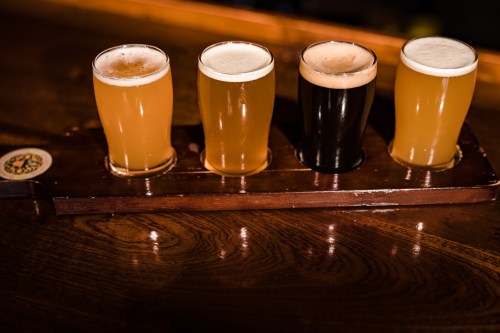 A wooden flight board holds four glasses of different beers: two pale, one amber, and one dark stout.