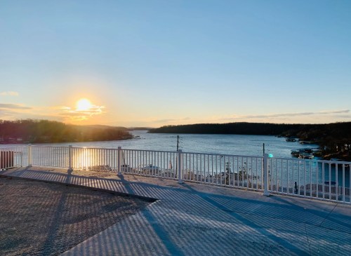 Sunset over a calm lake, with a white railing in the foreground and trees lining the distant shore.