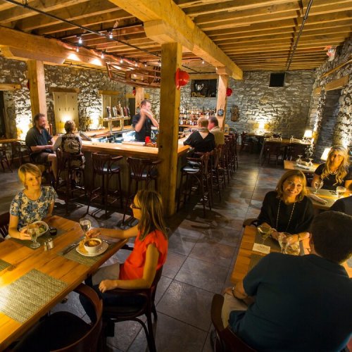 A cozy restaurant interior with stone walls, wooden beams, and patrons enjoying meals and drinks at tables and the bar.