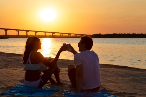 A couple toasting drinks at sunset on the beach, with a bridge in the background and water reflecting the golden light.