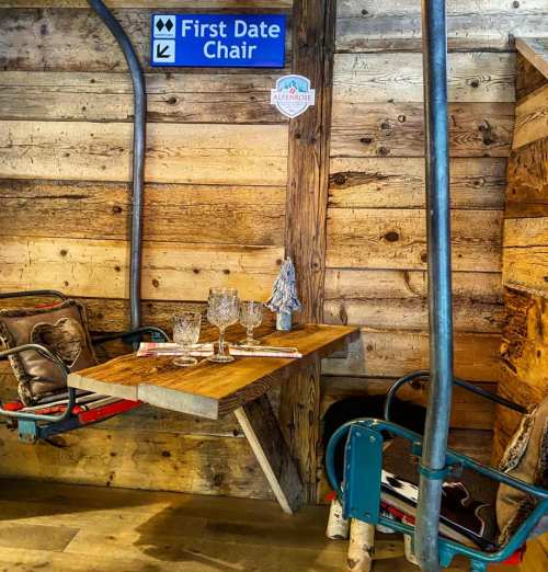 A rustic wooden table set for two, with glasses and utensils, beside a sign for "First Date Chair" in a cozy setting.
