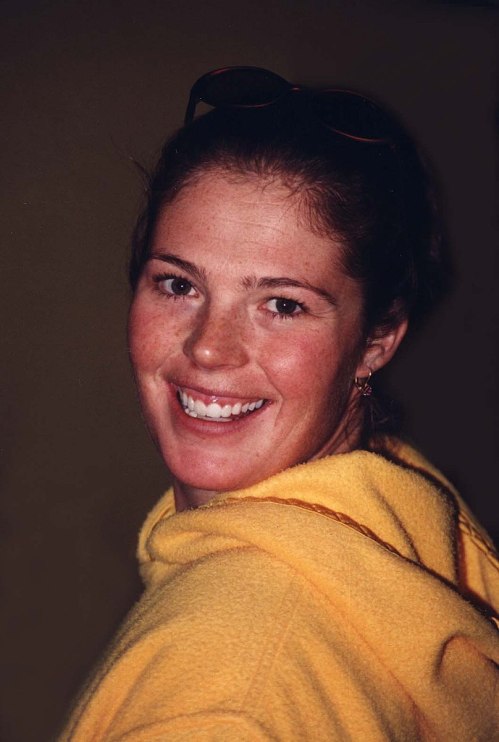 Smiling young woman in a yellow hoodie, with sunglasses on her head, looking over her shoulder.