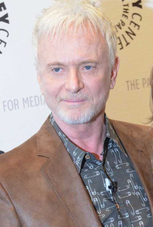 A man with short white hair and a beard, wearing a brown jacket and patterned shirt, smiles at an event.