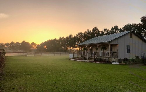 A serene sunrise over a rustic house surrounded by green grass and trees, with a fence in the background.