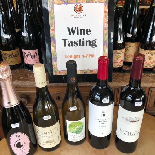 A sign for a wine tasting event with five bottles of wine displayed on a table. Event time: 4-7 PM.
