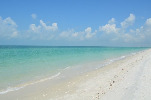 A serene beach with soft white sand, clear turquoise water, and a bright blue sky dotted with fluffy clouds.