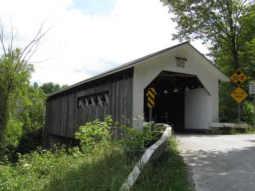 A rustic covered bridge surrounded by greenery, with a warning sign for height restrictions.