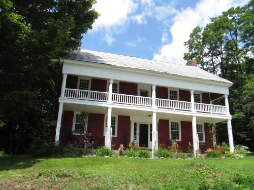 A red two-story house with white columns and a porch, surrounded by greenery and colorful flowers under a blue sky.