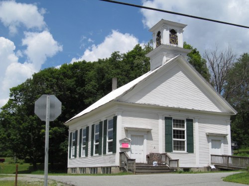 A white, historic building with green shutters, a bell tower, and a stop sign in front, surrounded by trees and blue sky.