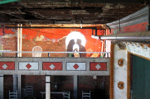 Two silhouettes are seen through a circular window in a rustic, partially abandoned interior with exposed brick and peeling paint.