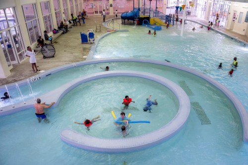 Aerial view of a busy indoor pool with children swimming, playing, and adults supervising in a bright, spacious environment.