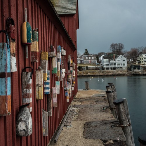 Colorful buoys hang on a red building by a calm harbor, with houses visible across the water under a cloudy sky.