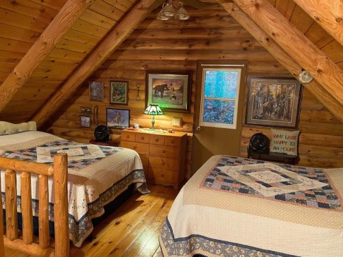 Cozy attic bedroom with two beds, wooden walls, and rustic decor, featuring paintings and a dresser.