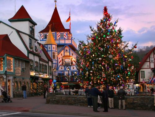 A festive town square with a large decorated Christmas tree and colorful buildings, as people gather in the evening light.