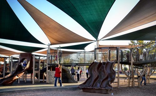 A playground with large shaded canopies, featuring slides and climbing structures, with children playing.