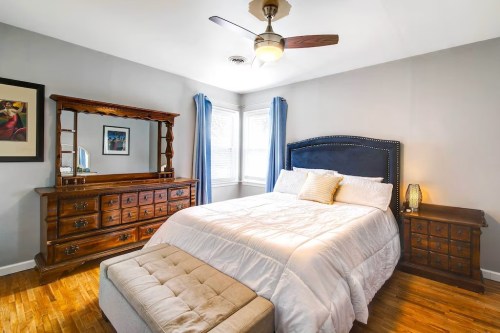 Cozy bedroom featuring a queen bed, wooden dresser, nightstand, and large windows with blue curtains.