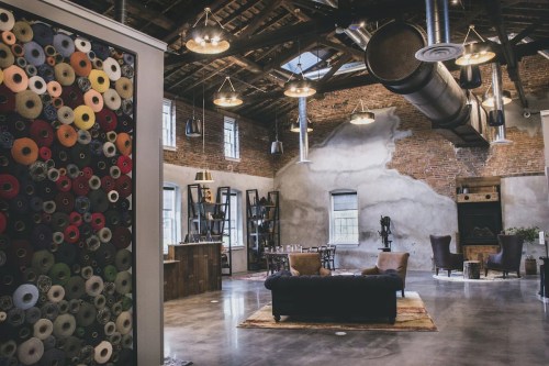 A spacious, modern loft with exposed brick walls, industrial lighting, and colorful wall art. Cozy seating area visible.