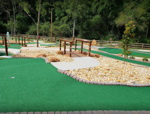 A scenic mini-golf course featuring green turf, wooden bridges, and landscaped areas with rocks and plants.