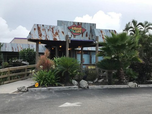 Exterior of a rustic restaurant named "Squid Lips," surrounded by tropical plants and a cloudy sky.