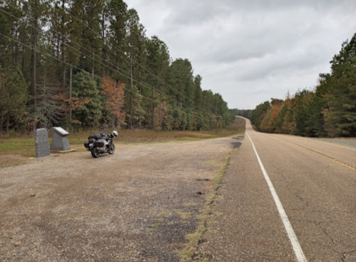 A motorcycle parked on a gravel area beside a winding road, surrounded by trees with autumn foliage.