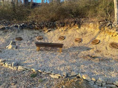 A wooden bench sits on a gravel area surrounded by tree stumps and a sloped, rocky landscape.