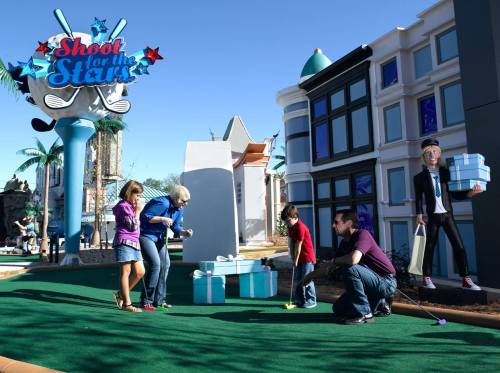A family plays mini-golf near a colorful "Shoot for the Stars" sign, with oversized gifts and a cartoon figure nearby.