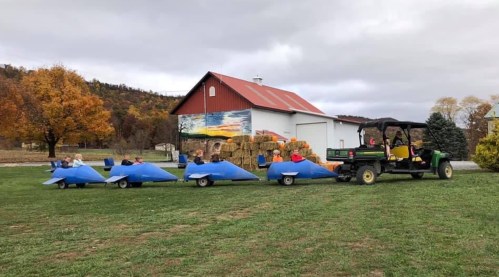 A tractor pulls a train of children in blue hay bales on a grassy field near a barn with autumn trees in the background.