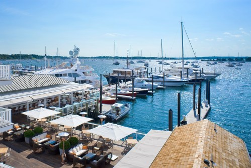 A sunny marina scene with yachts docked, a waterfront restaurant, and clear blue skies.