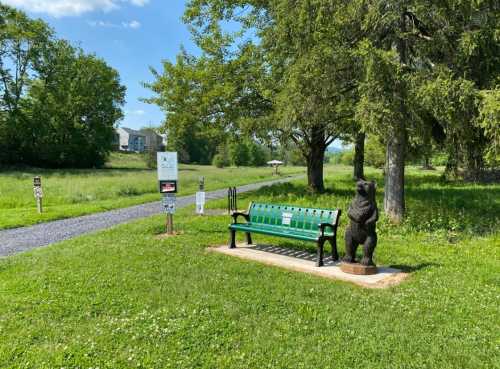 A green bench beside a bear statue in a grassy park with trees and a walking path in the background.