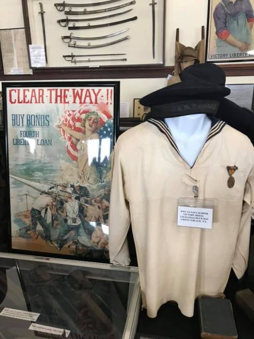 A vintage poster promoting the Fourth Liberty Loan beside a sailor's uniform and medals in a display case.