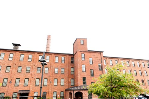 Historic brick building with multiple windows and a tall chimney, surrounded by greenery under a cloudy sky.