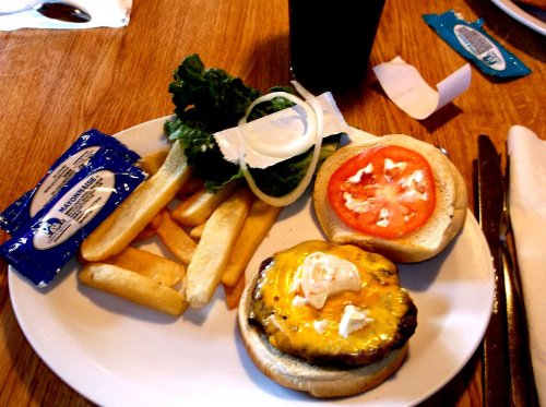 A plate with a cheeseburger, lettuce, tomato, onion, fries, and a packet of mayonnaise on a wooden table.