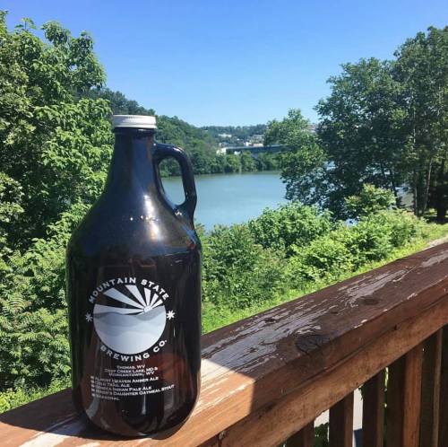 A glass growler from Mountain State Brewing Co. sits on a wooden railing with a scenic river and trees in the background.