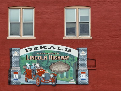 Sign for the Lincoln Highway in DeKalb, featuring a vintage car and historical imagery, against a red brick wall.