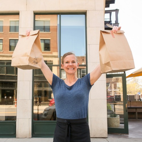 A smiling woman holds up two takeout bags outside a restaurant, showcasing her excitement.