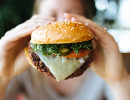 A person holds a large burger with lettuce, tomato, pickles, and melted cheese, with a blurred background.