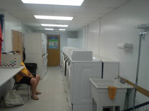 A laundry room with white machines, a sink, and a person sitting on a chair. Brightly lit and organized space.