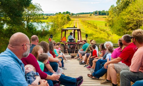 A group of people, including children, sit on a hayride, enjoying a scenic view of fields and trees.