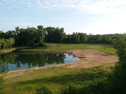 A serene landscape featuring a calm pond surrounded by green trees and a sandy shore under a clear blue sky.