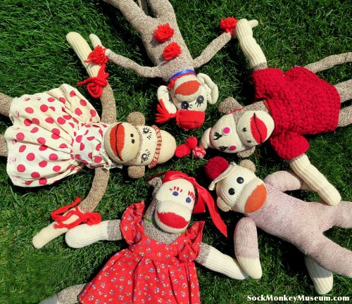 A circle of colorful sock monkeys in various outfits, lying on green grass, showcasing their playful expressions.