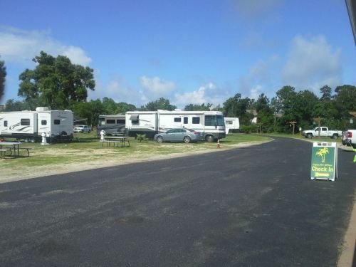 A campground with several RVs parked, picnic tables, and a "Check In" sign under a clear blue sky.