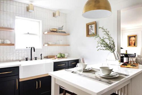 Modern kitchen with black cabinets, white sink, wooden shelves, and a dining table set for two, featuring a vase and wine.