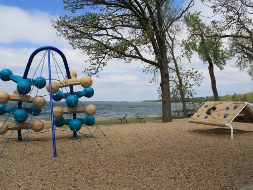 Playground with climbing structures by a lake, surrounded by trees and a cloudy sky.