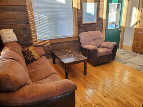 Cozy living room with a brown couch, armchair, marble coffee table, and wooden walls, featuring a green door and sunlight.